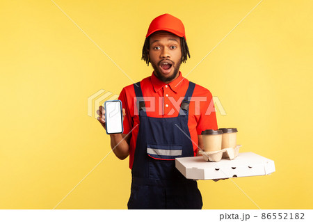 Portrait of surprised astonished courier man with open mouth wearing blue overalls holding smartphone with blank screen, pizza and take away coffee. Indoor studio shot isolated on yellow background. 86552182