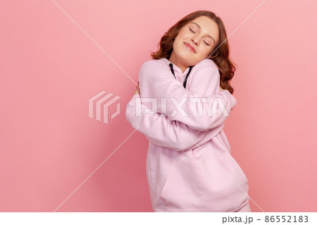 Portrait of selfish narcissistic brunette teen female in hoodie hugging herself and smiling with dreamy satisfied expression, love myself gesture. Indoor studio shot isolated on pink background 86552183