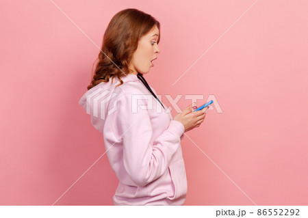 Profile of amazed curly haired teenage girl looking at cellphone with surprised expression, reading shocking news using mobile phone. Indoor studio shot isolated on pink background Profile of amazed curly haired teenage girl looking at cellphone with surprised expression, reading shocking news using mobile phone. Indoor studio shot isolated on pink background 86552292