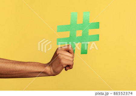 Social media concept. Closeup of human hand holding and showing large big green hashtag sign, copyspace. Indoor studio shot isolated on yellow background. 86552374