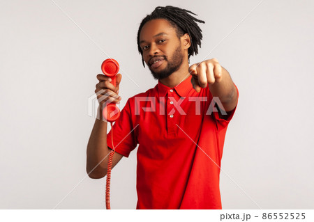 Handsome guy man with dreadlocks wearing red T-shirt, pointing finger to camera, holding handset of red vintage landline phone, answering calls. Indoor studio shot isolated on gray background. Handsome guy man with dreadlocks wearing red T-shirt, pointing finger to camera, holding handset of red vintage landline phone, answering calls. Indoor studio shot isolated on gray background. 86552525