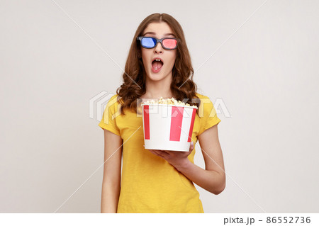 Astonished young female in 3d imax glasses watching movie film, hold popcorn, watching amazing movie, keeping mouth open, wears yellow T-shirt. Indoor studio shot isolated on gray background. Astonished young female in 3d imax glasses watching movie film, hold popcorn, watching amazing movie, keeping mouth open, wears yellow T-shirt. Indoor studio shot isolated on gray background. 86552736