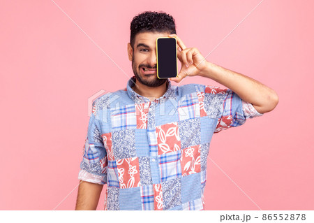 Portrait of funny bearded man wearing blue casual style shirt standing covering half face with phone, fooling around, making grimace. Indoor studio shirt isolated on pink background. Portrait of funny bearded man wearing blue casual style shirt standing covering half face with phone, fooling around, making grimace. Indoor studio shirt isolated on pink background. 86552878