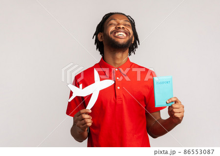 Extremely happy man with dreadlocks wearing red casual style T-shirt, holding passport document and paper airplane, rejoicing travel tour. Indoor studio shot isolated on gray background. Extremely happy man with dreadlocks wearing red casual style T-shirt, holding passport document and paper airplane, rejoicing travel tour. Indoor studio shot isolated on gray background. 86553087