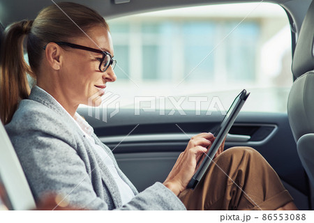 Side of a successful smiling business woman wearing eyeglasses using digital tablet, working while sitting on back seat in the car Side of a successful smiling business woman wearing eyeglasses using digital tablet, working while sitting on back seat in the car 86553088