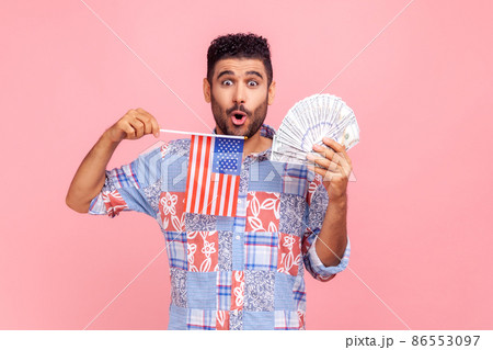 Astonished attractive man with beard and dark hair holding USA flag and dollars bills, looking at camera with open mouth, expressing shock. Indoor studio shot isolated on pink background. Astonished attractive man with beard and dark hair holding USA flag and dollars bills, looking at camera with open mouth, expressing shock. Indoor studio shot isolated on pink background. 86553097