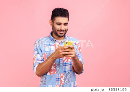 Portrait of attractive bearded man in blue shirt using mobile phone with happy expression, addicted to smartphone, texting in social network. Indoor studio shot isolated on pink background. Portrait of attractive bearded man in blue shirt using mobile phone with happy expression, addicted to smartphone, texting in social network. Indoor studio shot isolated on pink background. 86553196