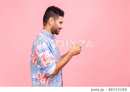 Side view of handsome dark haired man with beard with wearing casual style shirt using online service on mobile phone and expressing pleasant emotions. Indoor studio shot isolated on pink background. 86553269