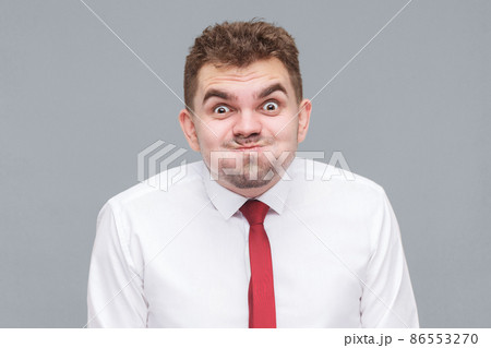 funny portrait of young man in white shirt and tie standing blowing his cheeks and looking at camera with funny face and big eyes. indoor isolated on gray background. 86553270