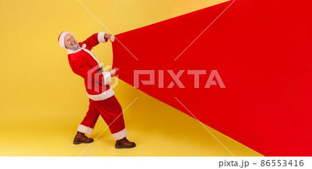 Extremely happy smiling elderly man with gray beard wearing santa claus costume pulling huge bag of gifts, copy space for advertisement. Indoor studio shot isolated on yellow background. 86553416