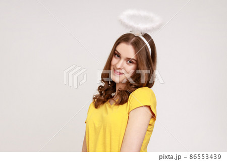 Shy attractive teenager girl with wavy hair in casual yellow T-shirt, standing with halo on her head and looking at camera and dreaming face. Indoor studio shot isolated on gray background. 86553439