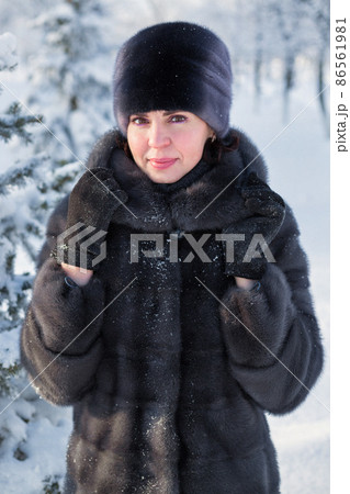 A woman in a mink fur coat and a hat with a smile stands against the background of fir trees covered with fluffy snow 86561981