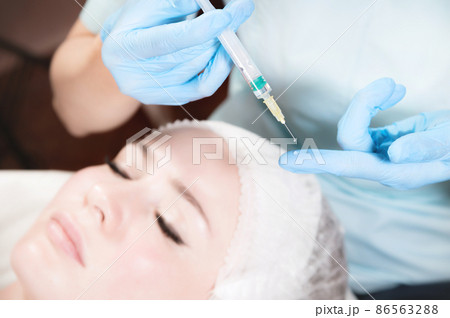 Close-up of a syringe with a drop of botox in the hand of a beautician against the background of the face of a caucasian girl. Injections and injectionology Close-up of a syringe with a drop of botox in the hand of a beautician against the background of the face of a caucasian girl. Injections and injectionology 86563288