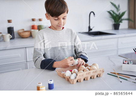 Cheerful concentrated child boy smiling and painting Easter egg at the kitchen 86565644