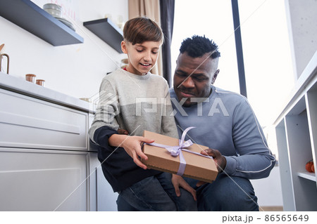 Child boy and his male parent hugging each other in the kitchen while looking at the gift box Child boy and his male parent hugging each other in the kitchen while looking at the gift box 86565649