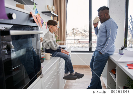 Boy sitting at the bartender and chatting with him multiracial father at the kitchen at the morning 86565843