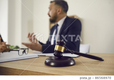 Close up of judge's gavel on wooden table, with lawyer giving consultation in background, explaining controversial case, providing professional legal advice on problem in court. Law services concept Close up of judge's gavel on wooden table, with lawyer giving consultation in background, explaining controversial case, providing professional legal advice on problem in court. Law services concept 86568970