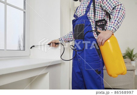 Pest control guy spraying poisonous gas or liquid on a windowsill at home. Male exterminator in overall uniform workwear spraying insecticide over a modern white window and sill inside the house 86568997