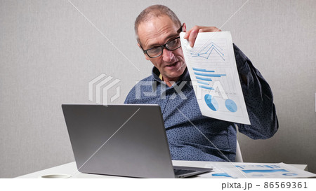 Elderly man checks drawings near laptop at white office desk 86569361