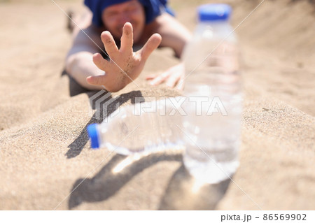 A thirsty man lies on the sand reaching for a bottle of water 86569902