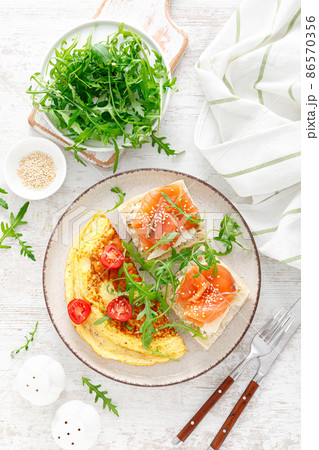 Omelette or omelet, fresh arugula and tomato salad and toasts with butter and salted salmon. Breakfast. Top view Omelette or omelet, fresh arugula and tomato salad and toasts with butter and salted salmon. Breakfast. Top view 86570356