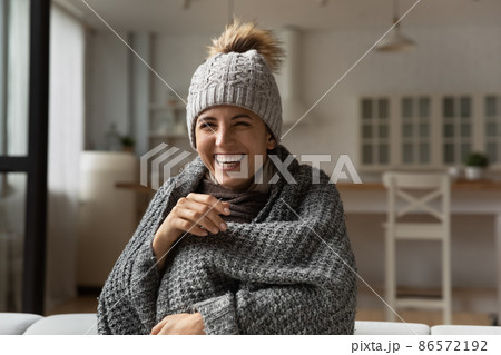 Laughing woman wear hat wrapped in knitted plaid sit indoor 86572192