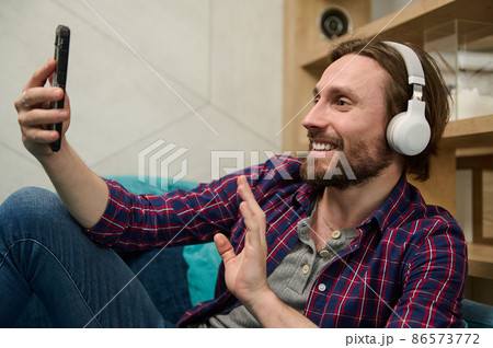 Handsome man having fun discussion by video call, wearing wireless headphones and smiles looking at a smartphone web camera, greeting and waving hello while relaxing at home Handsome man having fun discussion by video call, wearing wireless headphones and smiles looking at a smartphone web camera, greeting and waving hello while relaxing at home 86573772