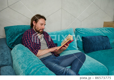 Serene confident young bearded Caucasian man relaxing on the couch, reading book in home environment. Education, study, knowledge and erudition concept 86573775