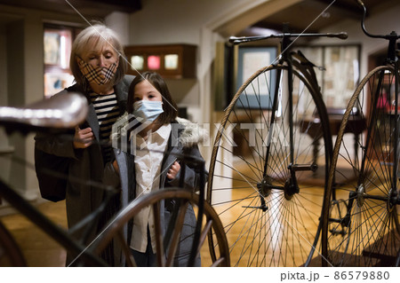 Tween schoolgirl and elderly female tutor in face masks viewing vintage bicycle in museum 86579880