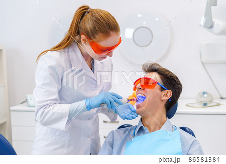 Female doctor in medical uniform with latex gloves and protective goggles putting curing light into opened mouth of young man during work in modern dental clinic 86581384