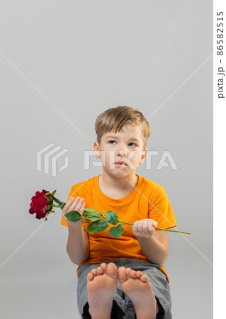 adorable preschool sitting barefoot. He is looking at camera while holding rose. On grey background. adorable preschool sitting barefoot. He is looking at camera while holding rose. On grey background. 86582515