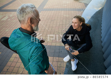 High angle view of sportive mature couple, man and woman in sportswear talking outdoors while waiting together for workout High angle view of sportive mature couple, man and woman in sportswear talking outdoors while waiting together for workout 86585574