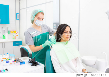 The dentist's assistant puts the protection on the patient, the napkin on the chest The dentist's assistant puts the protection on the patient, the napkin on the chest 86586163