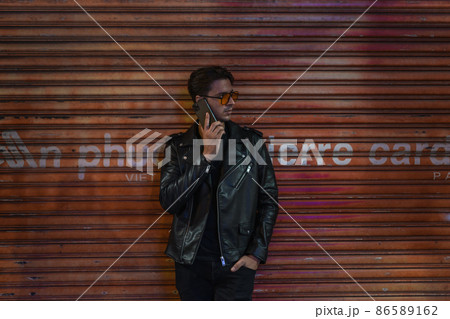 Portrait of young stylish brunet caucasian man in black leather jacket and yellow sunglasses. Standing near the wall and talking by phone. 86589162