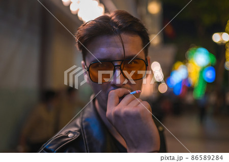 Close up portrait of young handsome brunet caucasian man in black leather jacket and yellow sunglasses. Standing on the street at night and smoking. Close up portrait of young handsome brunet caucasian man in black leather jacket and yellow sunglasses. Standing on the street at night and smoking. 86589284