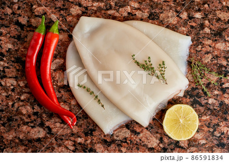 Three carcasses of fresh raw squid with lemon, pepper, thyme on a marble stone dark table. Three carcasses of fresh raw squid with lemon, pepper, thyme on a marble stone dark table. 86591834