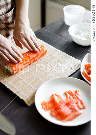 Young woman wrapping up sushi roll ingredients on bamboo mat. 86592148