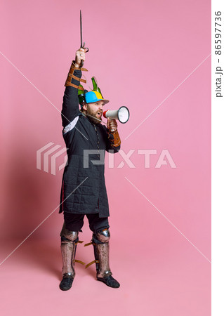 Full-length portrait of young man, medieval warrior or archer in beer helmet rising sword and shouting in megaphone isolated over pink background 86597736