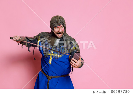 Half-length portrait of man, medieval warrior or knight in armor pointing sword at phone screen isolated over pink studio background 86597780