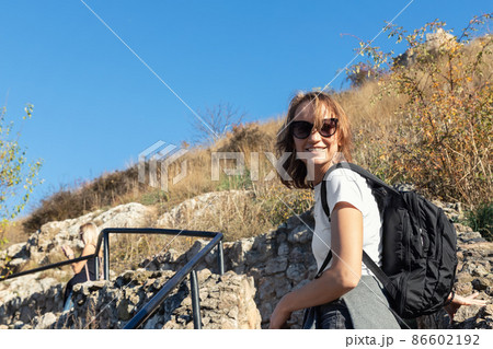 Young adult beautiful happy caucasian woman enjoy walking with backpack outdoors hill and mountain. Female tourist hiking outside at cliff staircase. Portrait smiling traveling active lady Young adult beautiful happy caucasian woman enjoy walking with backpack outdoors hill and mountain. Female tourist hiking outside at cliff staircase. Portrait smiling traveling active lady 86602192