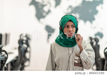Afro Muslim female with green hijab scarf customer representative business woman with phone headset helping and supporting online with customer in modern call centre Afro Muslim female with green hijab scarf customer representative business woman with phone headset helping and supporting online with customer in modern call centre 86603390