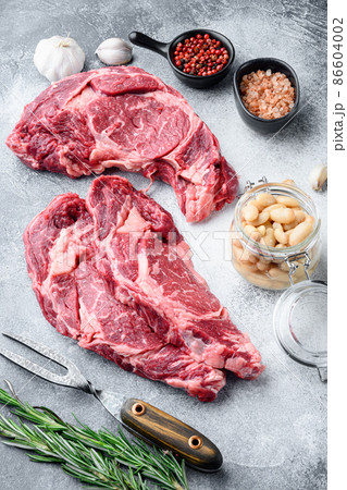 Raw rib eye beef marbled meat steak with pepper and herbs, on gray stone background Raw rib eye beef marbled meat steak with pepper and herbs, on gray stone background 86604002