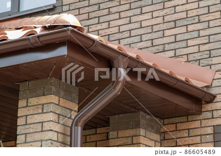 Corner of house with new brown metal tile roof and rain gutter. Metallic Guttering System, Guttering and Drainage Pipe Exterior 86605489