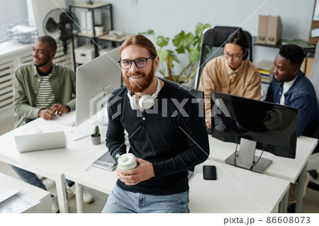 Happy young businessman with drink standing by desk with computer monitors Happy young businessman with drink standing by desk with computer monitors 86608073