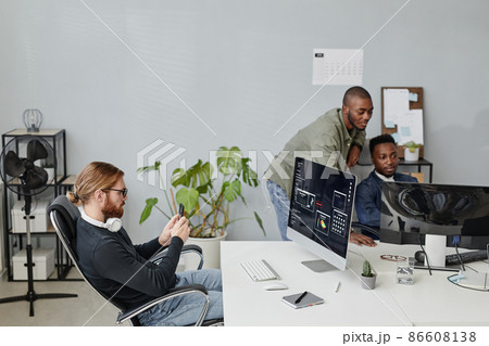 Young businessman with smartphone sitting by desk in front of computer screen Young businessman with smartphone sitting by desk in front of computer screen 86608138