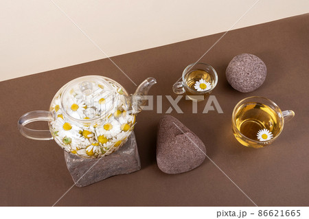 Chamomile tea. Cups of herbal tea, transparent teapot with camomile flowers and stones on brown background. Calming drink concept. Trendy still life Chamomile tea. Cups of herbal tea, transparent teapot with camomile flowers and stones on brown background. Calming drink concept. Trendy still life 86621665