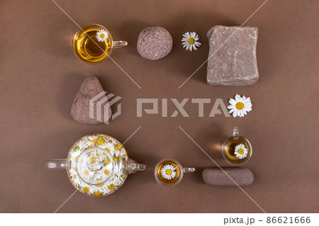 Chamomile tea. Cups of herbal tea, transparent teapot with camomile flowers and stones on brown background. Calming drink concept. Trendy still life Chamomile tea. Cups of herbal tea, transparent teapot with camomile flowers and stones on brown background. Calming drink concept. Trendy still life 86621666