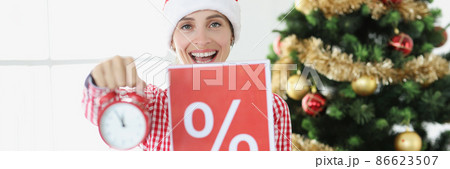 Young smiling woman in santa claus hat holds alarm clock and poster with percent on background of New Year tree Young smiling woman in santa claus hat holds alarm clock and poster with percent on background of New Year tree 86623507
