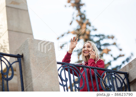 Attractive smiling woman talking on a cellphone and greeting someone with her hand in an autumn urban park outdoor, selective focus. Attractive smiling woman talking on a cellphone and greeting someone with her hand in an autumn urban park outdoor, selective focus. 86623790