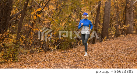 Caucasian adult woman in headset and blue sportswear is jogging in the forest in golden autumn, format image 1x2, selective focus Caucasian adult woman in headset and blue sportswear is jogging in the forest in golden autumn, format image 1x2, selective focus 86623885